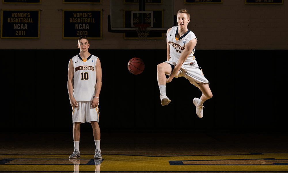 Two URochester basketball players on the court, one passing the other the basketball in the air underneath his legs