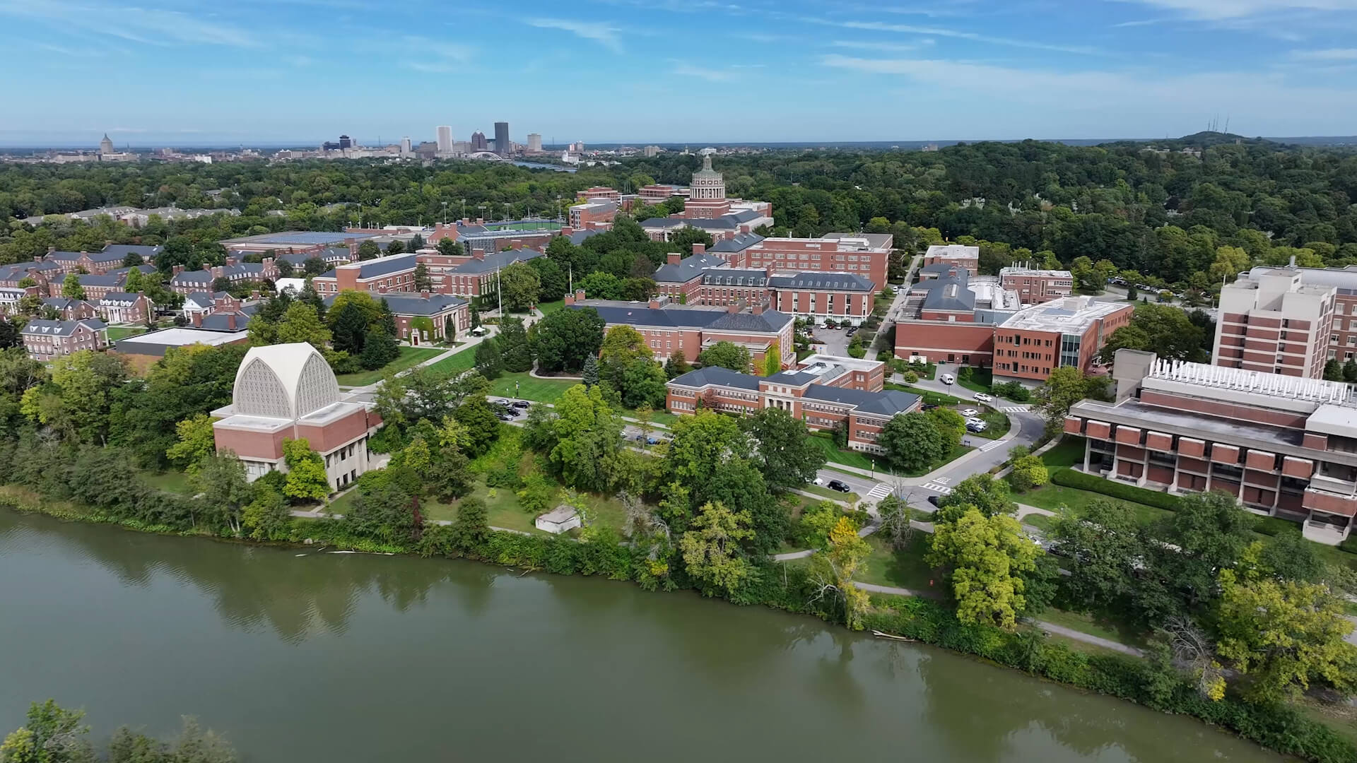 The Genesee River curves along Rochester’s leafy campus, where redbrick buildings rise from the green and the city skyline meets an open blue sky.