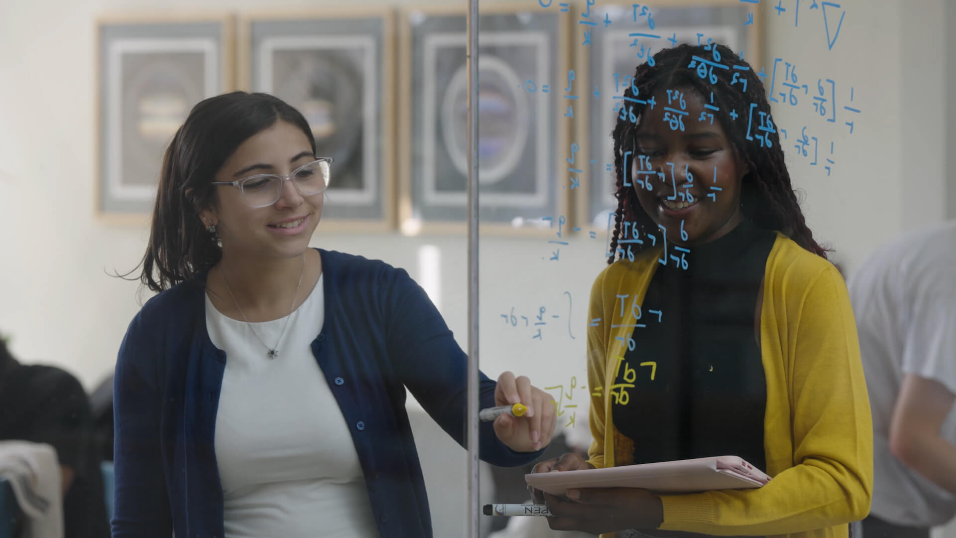 Two students share a smile over equations on a glass board, their ideas taking shape in blue and gold.