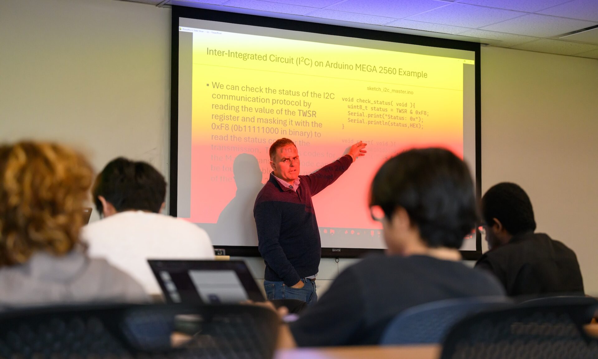 A URochester professor points to a projected lesson on computer programming and electronics while students follow along in a classroom.