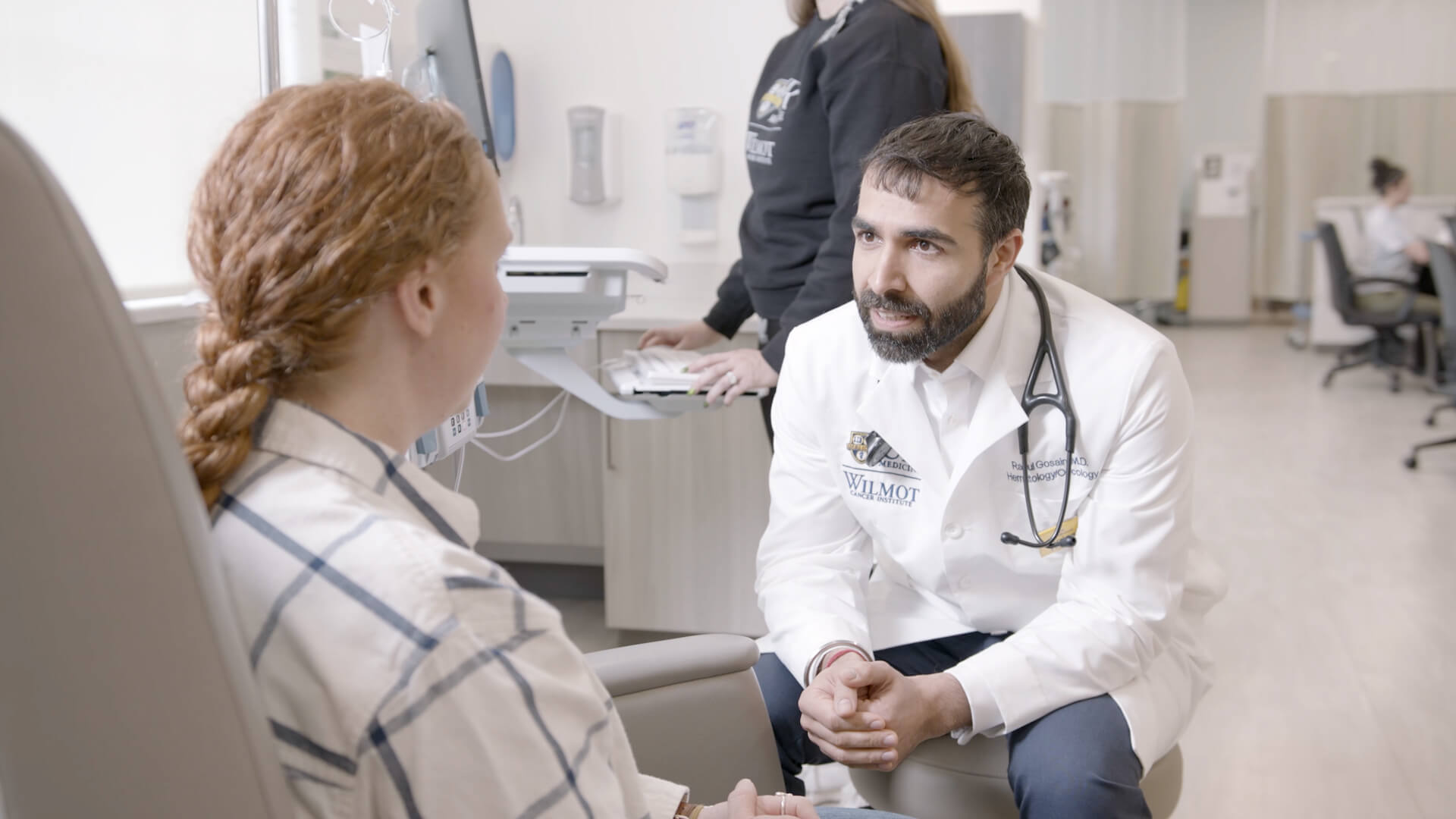 A physician leans close at a seated patient’s eye level, hands folded and attention steady in a quiet moment of human connection.