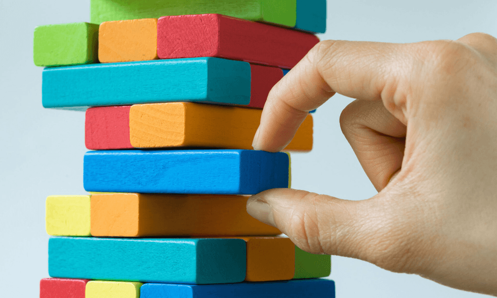Close-up of a hand pulling out a blue colored block from a Jenga-like stack of colorful blocks to illustrate the removal of the cGAS protein from cells.