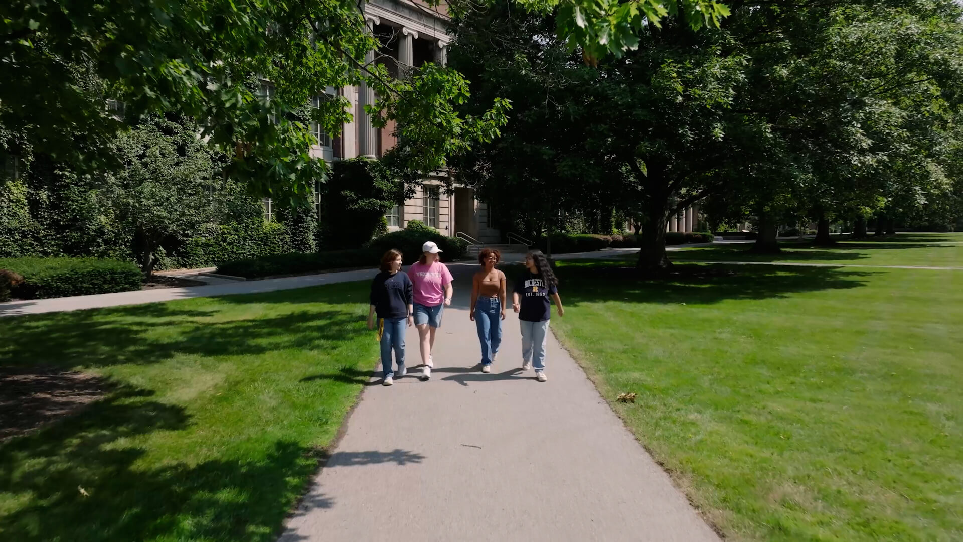 Four students walk side by side through URochester’s leafy campus, sharing conversation as sunlight and shade weave across their path.