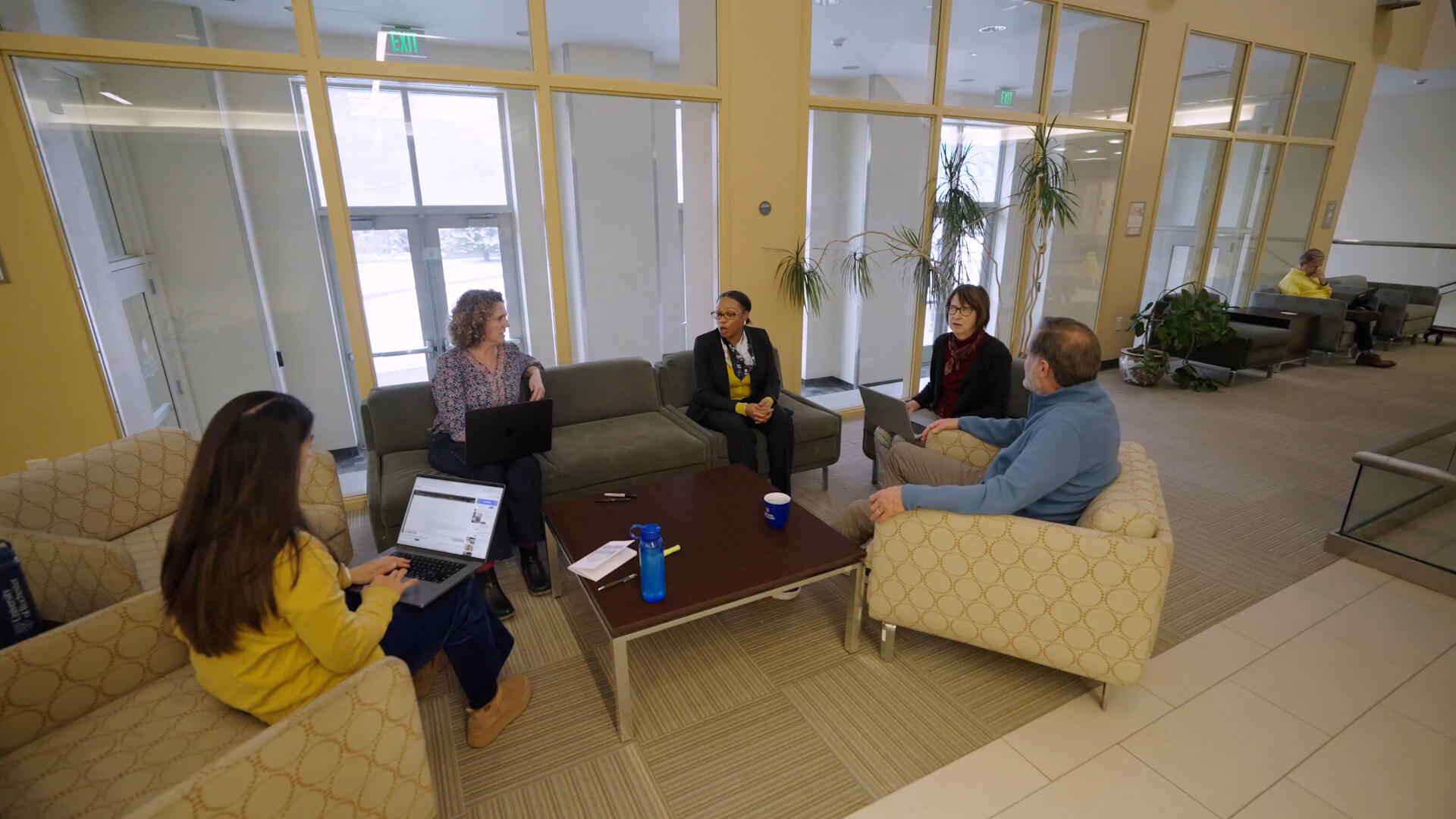 Five colleagues gather around a coffee table in a sunlit URochester lounge, laptops open and conversation bringing them together.