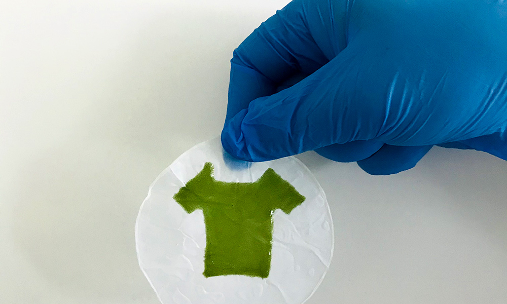 a blue-gloved hand holds a tiny circle of white material with a tiny green t-shirt printed on it.