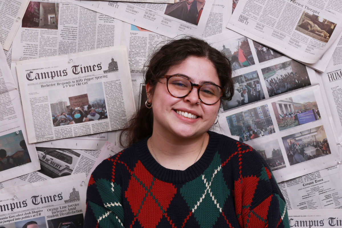 A smiling woman in glasses and a sweater stands before a pile of newspapers at the University of Rochester.