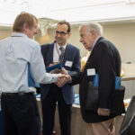Three professionals in business attire shake hands and converse during a networking event, with conference materials and attendees visible in the background.