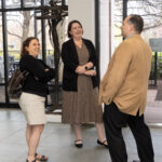 Two women and a man in business attire laugh and chat near a windowed entrance during a networking event.