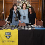 Three women pose in front of a check-in table with name tags and the University of Rochester banner displayed.