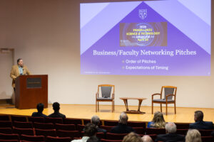 Picture of a stage with a middle aged man standing at a podium with a power point presentation on wall behind him with the words Business/Faculty Networking Pitches