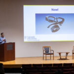 Barry Silverstein presenting at the podium during his Keynote talk with a powerpoint slide displayed on the wall - the slide has the word "Now!" at the top of it and contains picture of smart glasses and a smart band. The University of Rochester Center for Extended Reality wordmark is also displayed in the lower left corner.