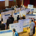 A zoomed out picture of an auditorium full of posters with numerous student and people interacting during the poster session.