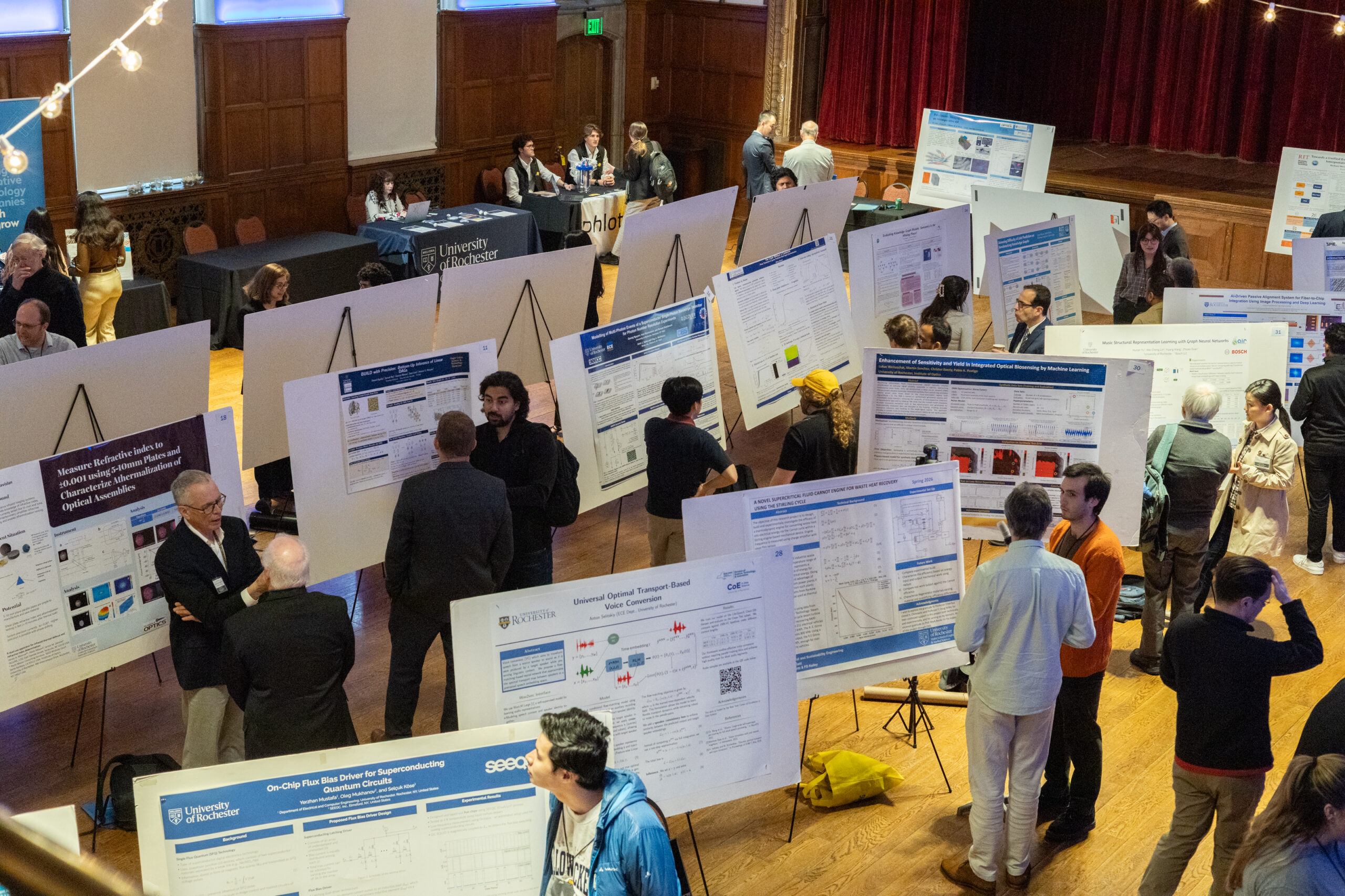 A zoomed out picture of an auditorium full of posters with numerous student and people interacting during the poster session.