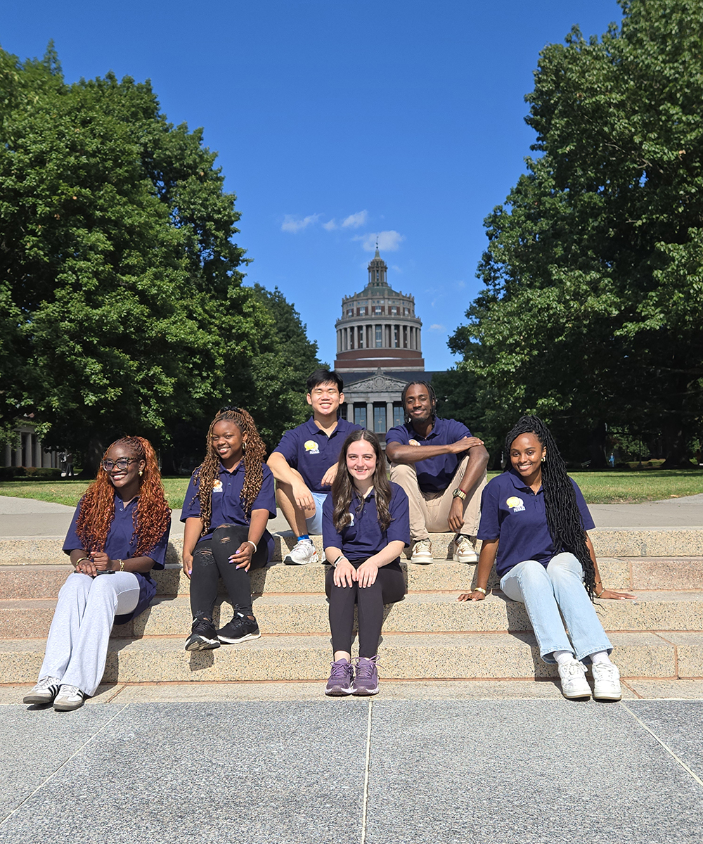 The 2025 peer advisors sitting on the steps to Eastman Quad with the tower or Rush Rhees in the background.