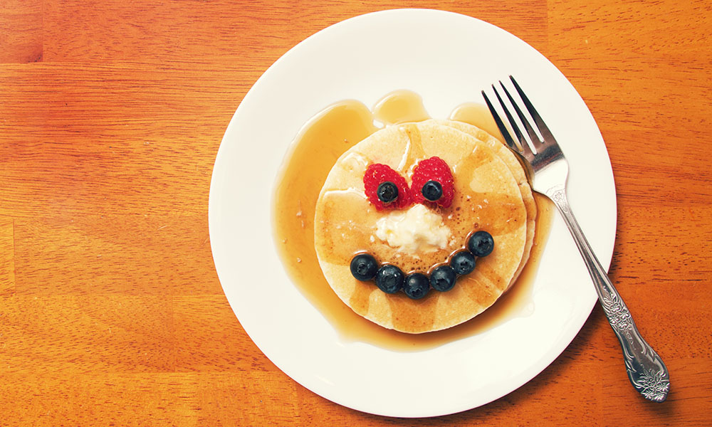 A syrup-covered pancake on a plate with blueberries for a smile and raspberries for eyes.