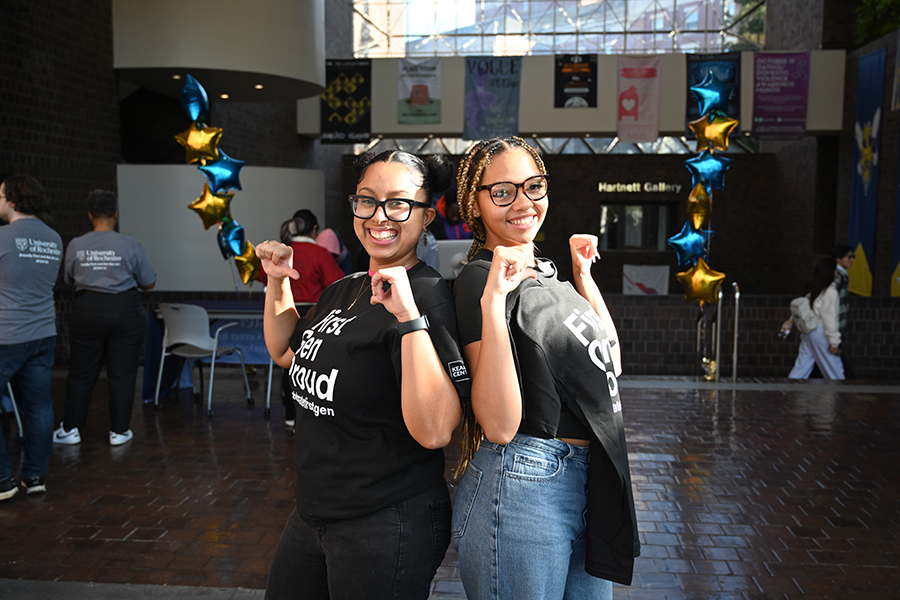 Two students wearing black t-shirts stand back to back pointing their thumbs at themselves.