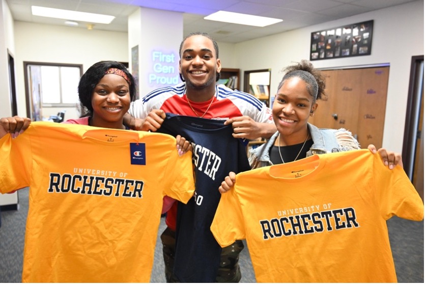 Three students smiling at the camera while holding up t-shirts.
