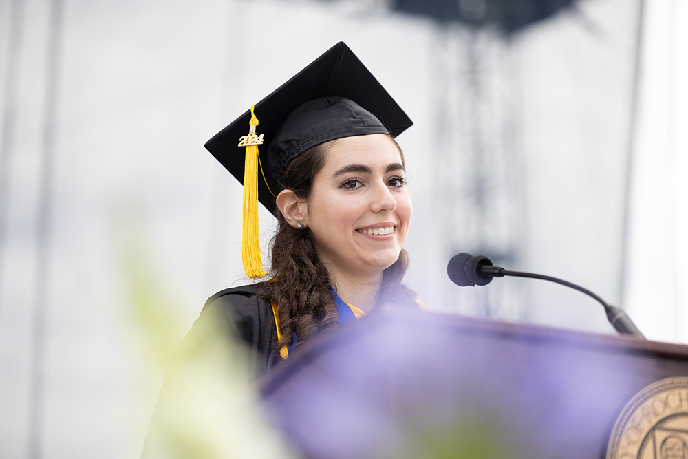 A previous student commencement speaker standing at a podium in a cap and gown.