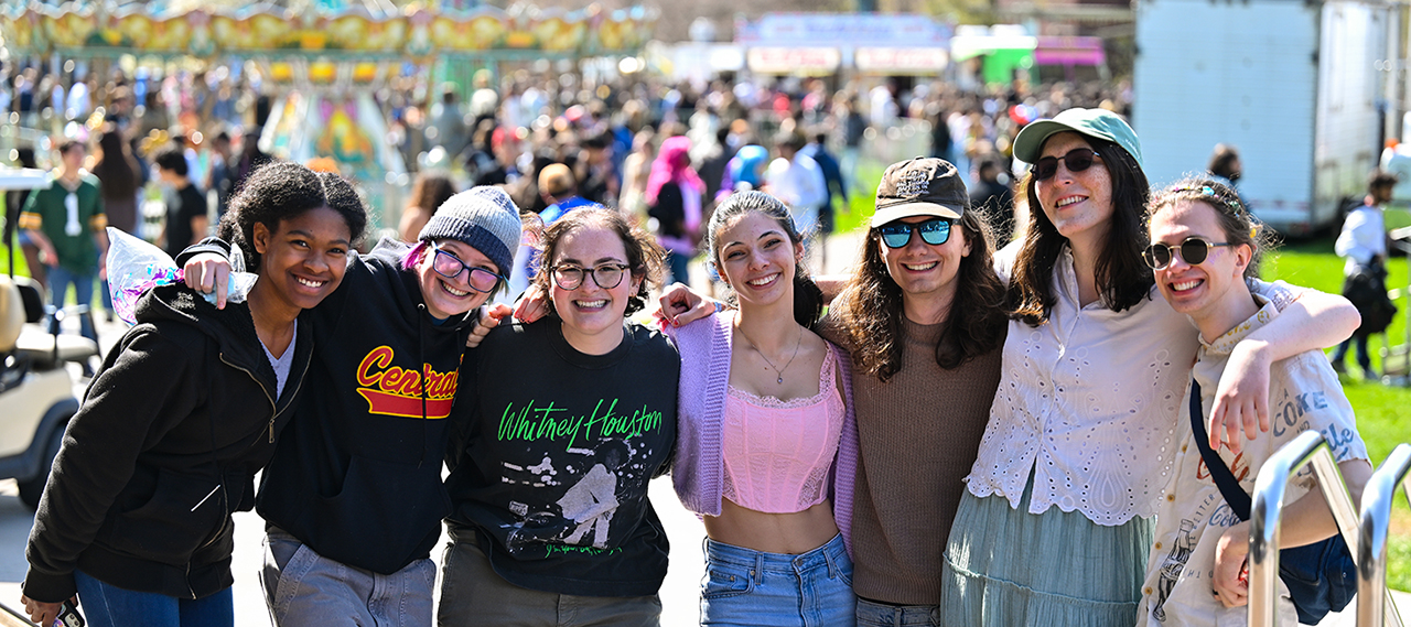 A group of students pose smiling looking at the camera outside on Wilson Quad during Dandelion Day with a carousel and group of students behind them.