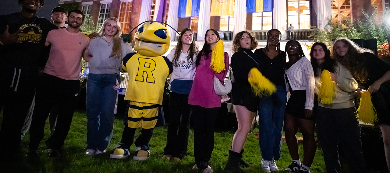 A group of students pose smiling looking at the camera in front of Rush Rhees Library at night with our mascot Rocky.