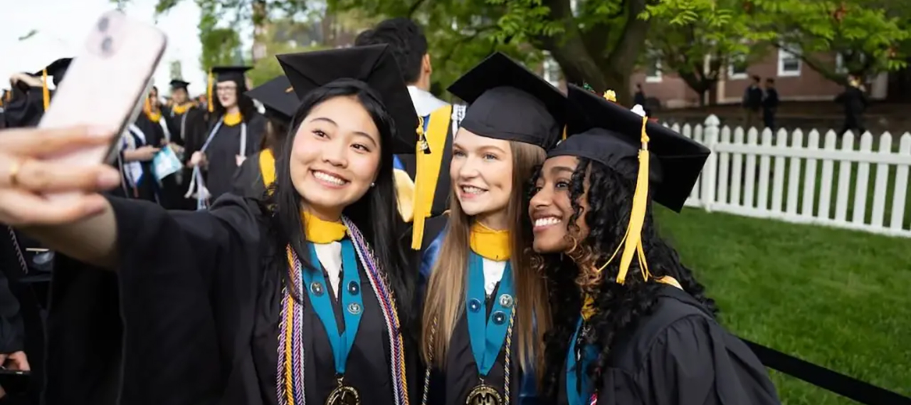 Three students taking a selfie outside during commencement in their regalia and wearing Medallion Leadership Program medallions.