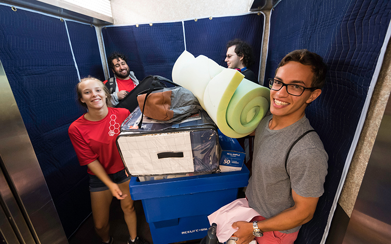 Photo of students entering the elevator in Susan B. Anthony Residence Halls carrying bins and a foam mattress pad