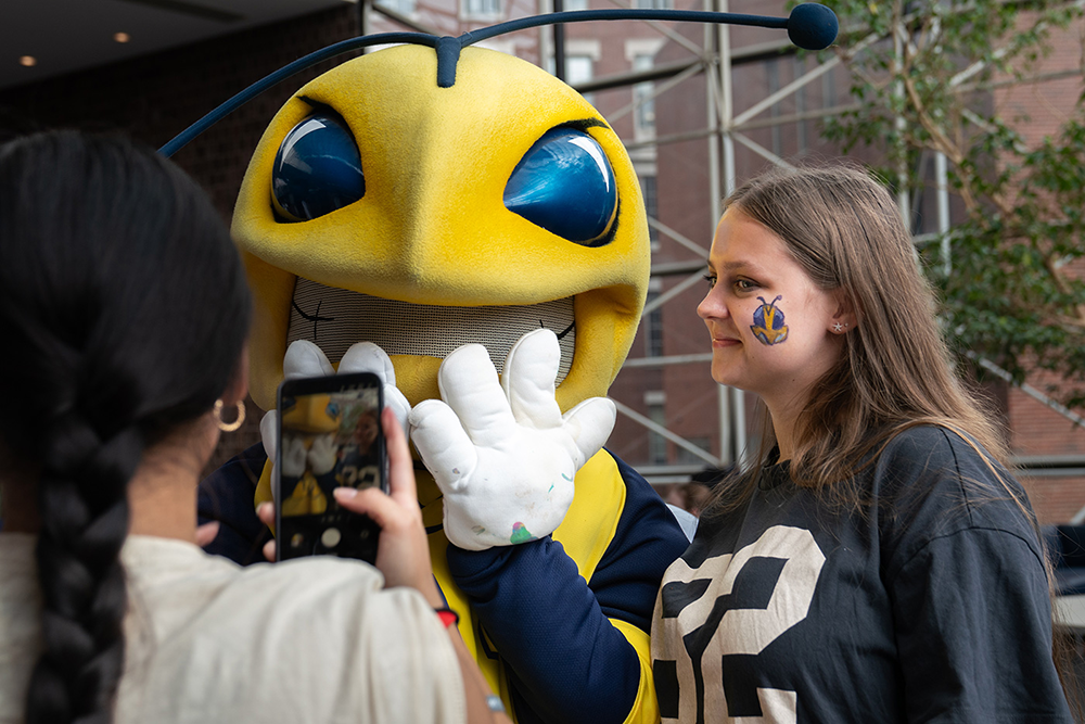 A student poses for a photo with Rocky while another student takes the photo with their phone in the foreground