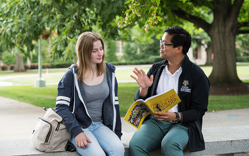 Photo of an advising staff member sitting and talking with a student on a bench on the Eastman Quad on River Campus