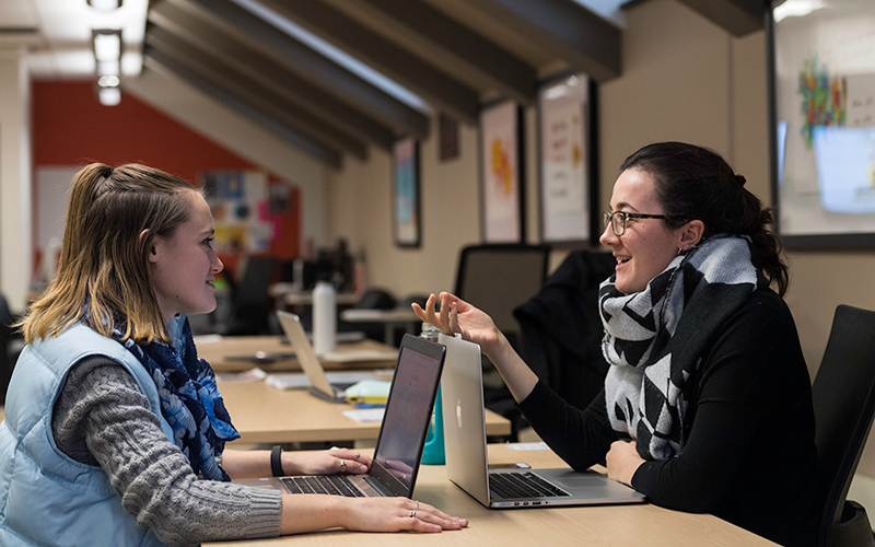 Photo of two people sitting across from one another talking with laptops in front of them
