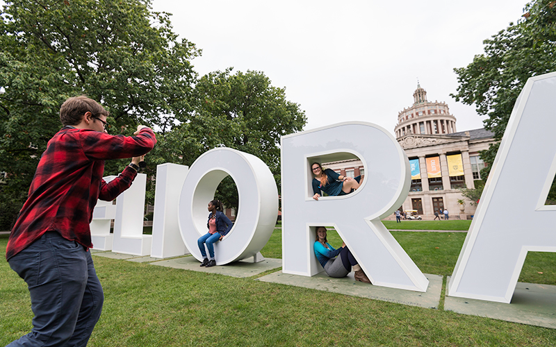 Photo of a person taking photos of other people posed inside the giant Meliora letters on the Eastman Quad during Meliora Weekend