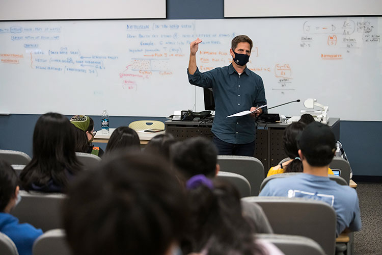 A professor teaching a class with a whiteboard in the background.