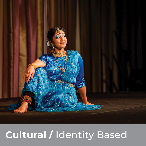 Cultural/Identity Based - Woman wearing colorful, traditional dress smirking in front of a stage curtain