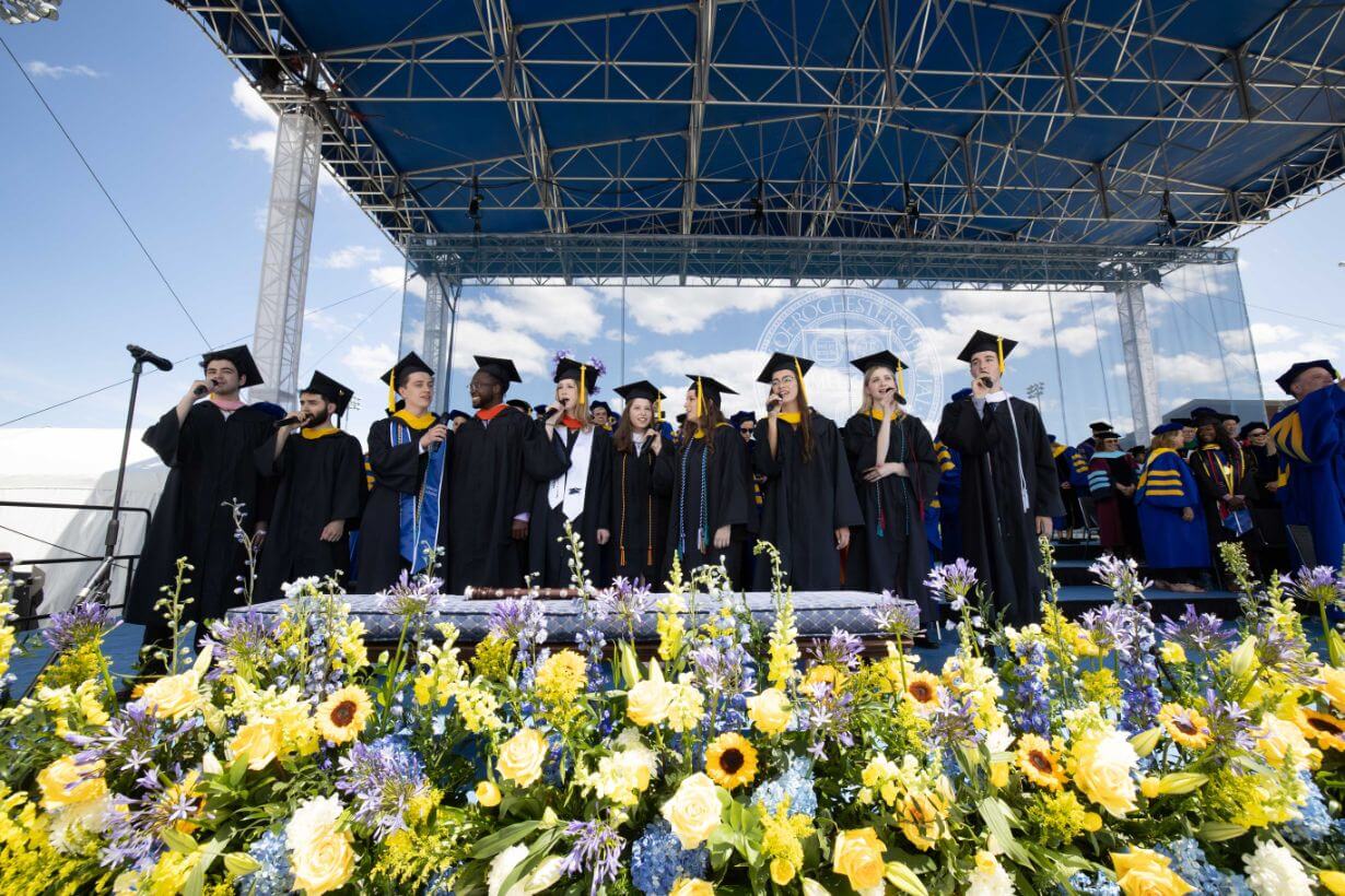 A group of graduates stands before a flower-decorated stage at the University of Rochester Commencement Weekend.