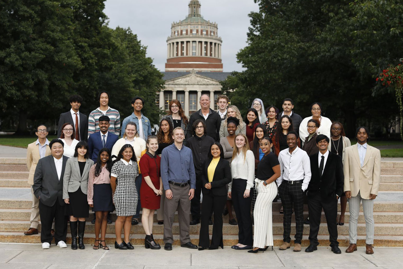 A group of students, along with Rich Handler, smile for a photo in front of the University of Rochester Eastman Quad, showcasing their camaraderie and enthusiasm.