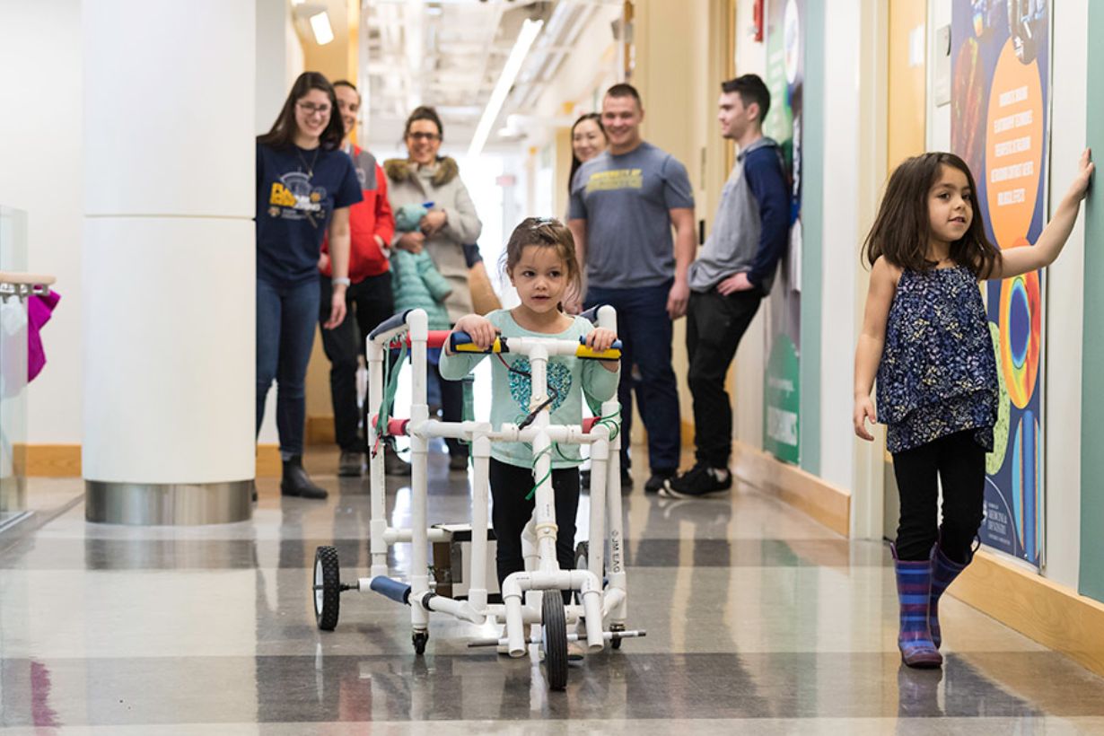 Four students watch a child walk in their walker prototype, with wheels and handlebars and plastic piping.
