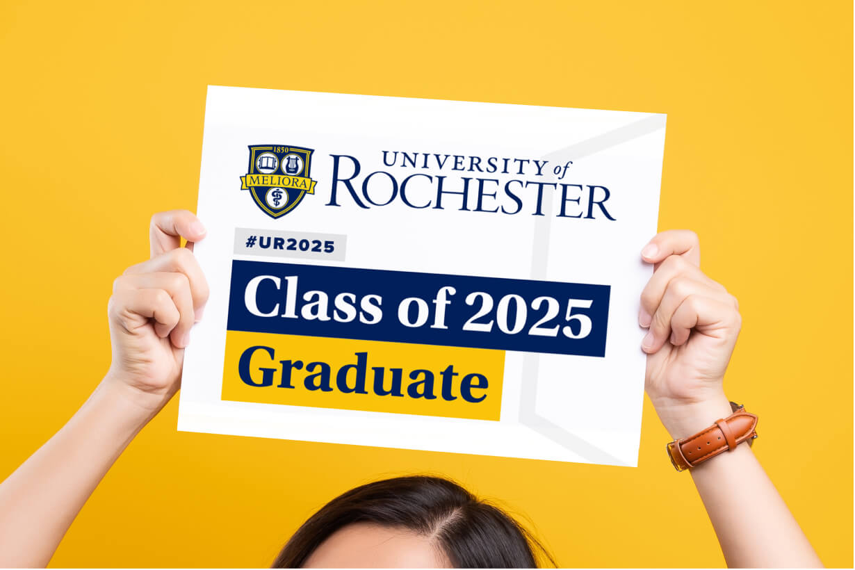 A University of Rochester graduate in cap and gown, celebrating their achievement with a sign in hand.