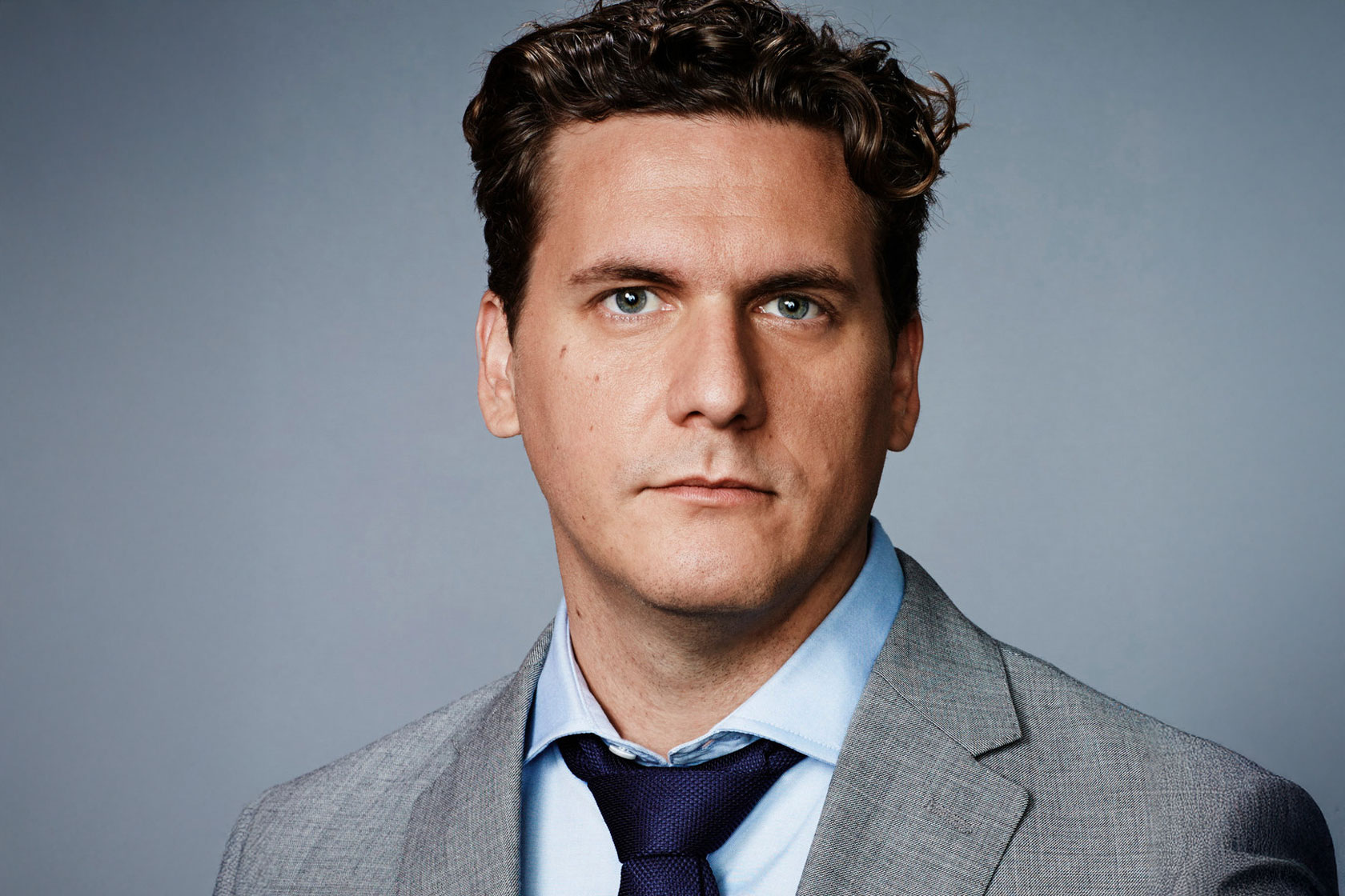 A man in a suit and tie gazes confidently at the camera, representing journalist Tommy Evans ’99 for the 2025 Commencement.