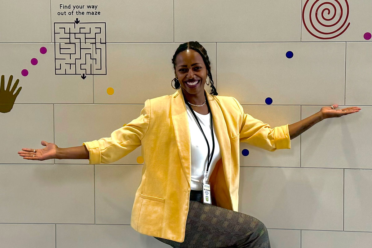 Katrina Grantham, a University of Rochester Class of 2025 student, stands in a yellow blazer before a colorful wall design.