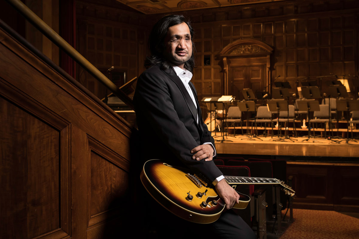 A man in a suit, Eshaan Sood, holds a guitar in front of an orchestra, representing the University of Rochester Class of 2025.