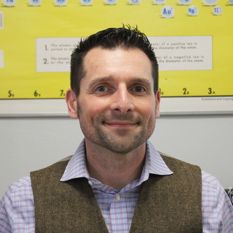 Jamie Porta, a chemistry teacher at Brighton High School, stands in front of a bright yellow wall wearing a vest and shirt.