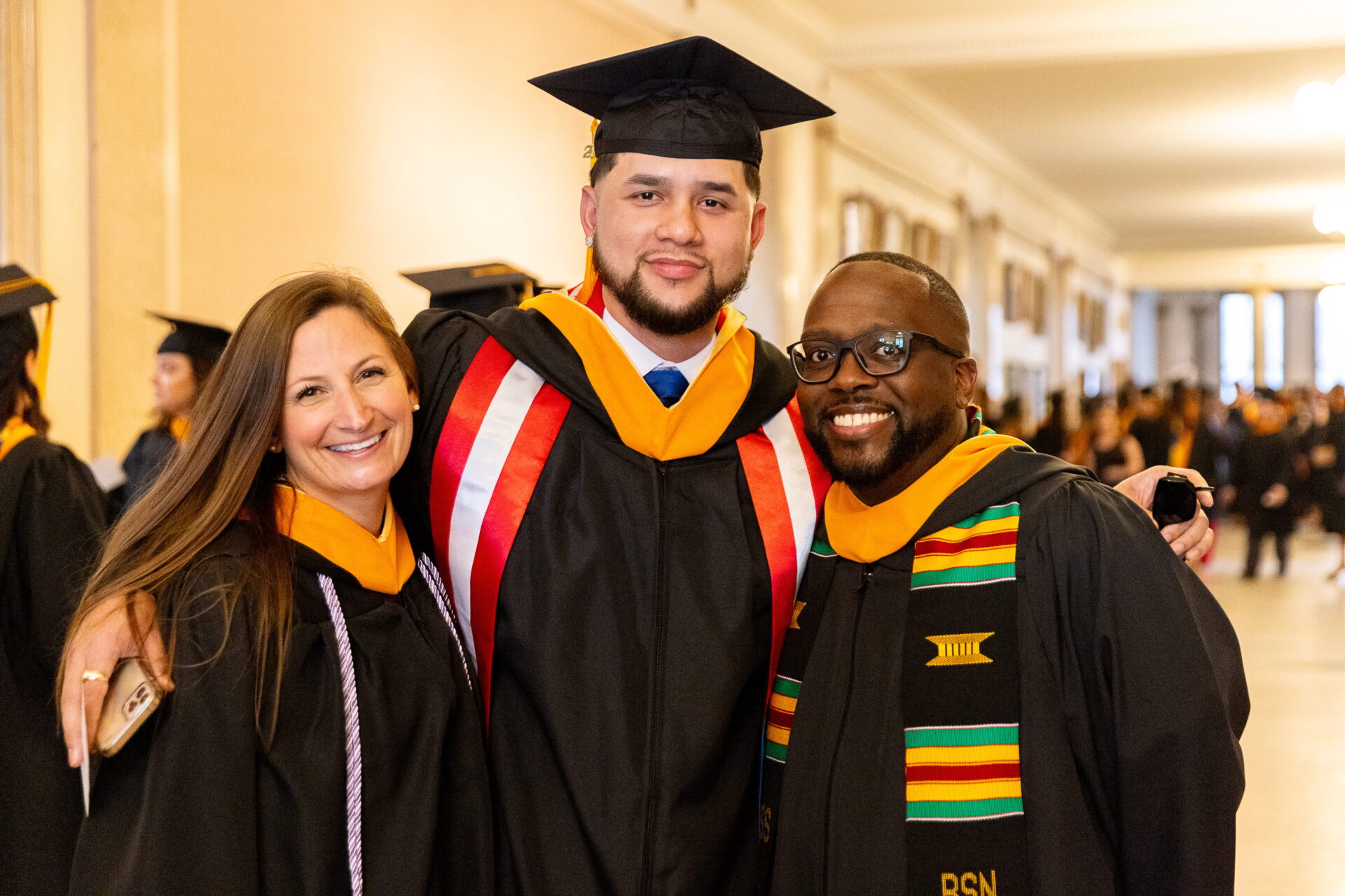 Three graduates smile together in a photo. Each is wearing a black cap and gown with a yellow stole.