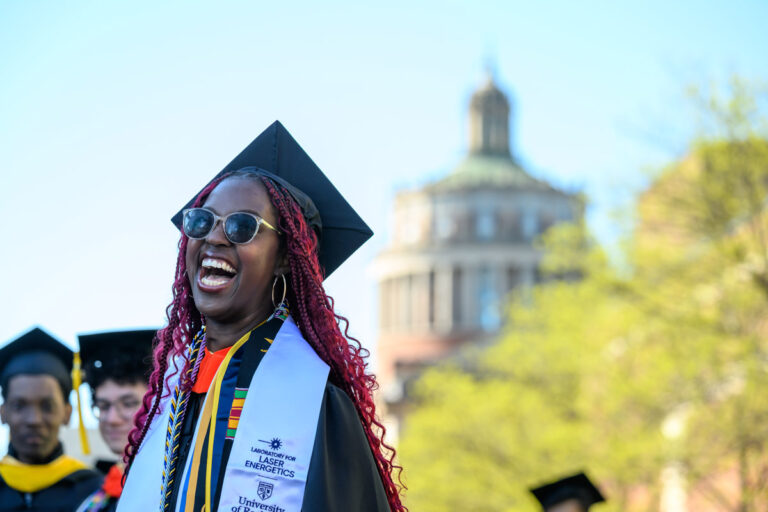 A University of Rochester graduate smiles during Commencement Weekend with Rush Rhees Library in the background.
