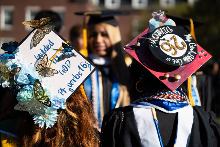 Two University of Rochester graduates wear decorated caps during Commencement Weekend, including one that reads Guided by God. Proverbs 16:9.