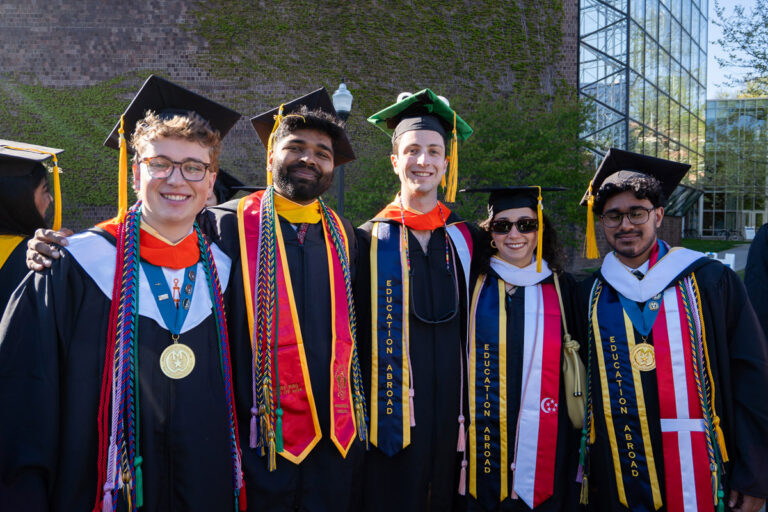 Five University of Rochester graduates pose together in decorated academic regalia during Commencement Weekend.