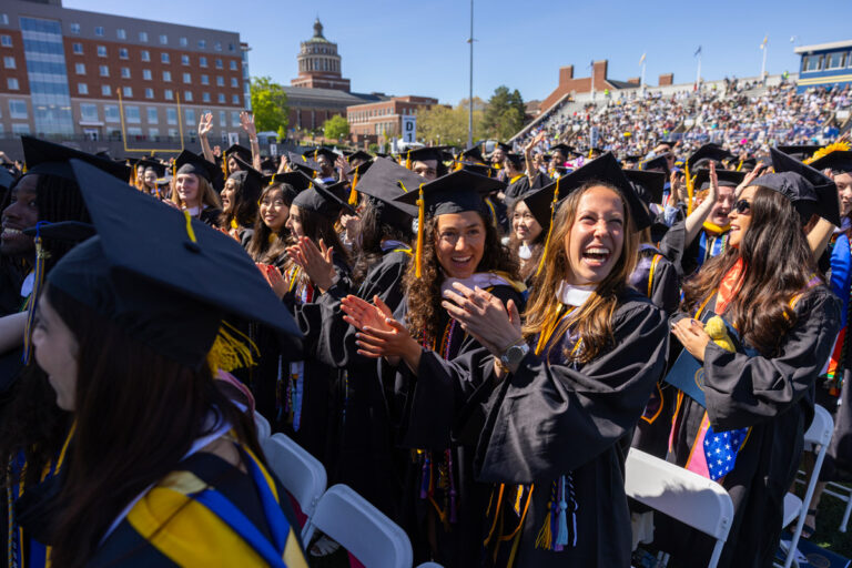 University of Rochester graduates smile and applaud during Commencement Weekend with Rush Rhees Library in the background.