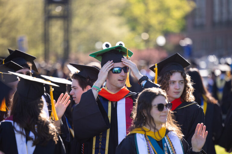 University of Rochester graduates process through campus, including one wearing a decorated frog-themed cap during Commencement Weekend.