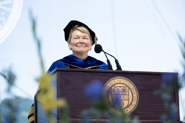 A University of Rochester speaker stands at the Commencement Weekend podium with flowers in the foreground.