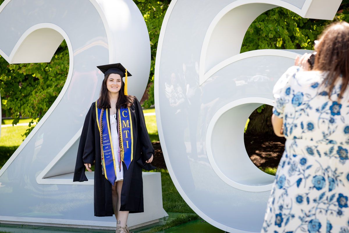 A woman in a graduation gown stands proudly next to a large 2026 sign at the University of Rochester.