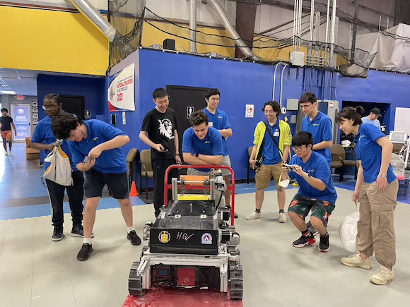 Engineering students in blue University of Rochester shirts surround a lunar rover they constructed