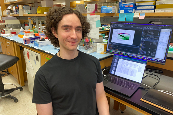 Alejandro Cruz Setzekorn smiles at the camera with a laptop and momitor in the background showing data science genomics research and figures.